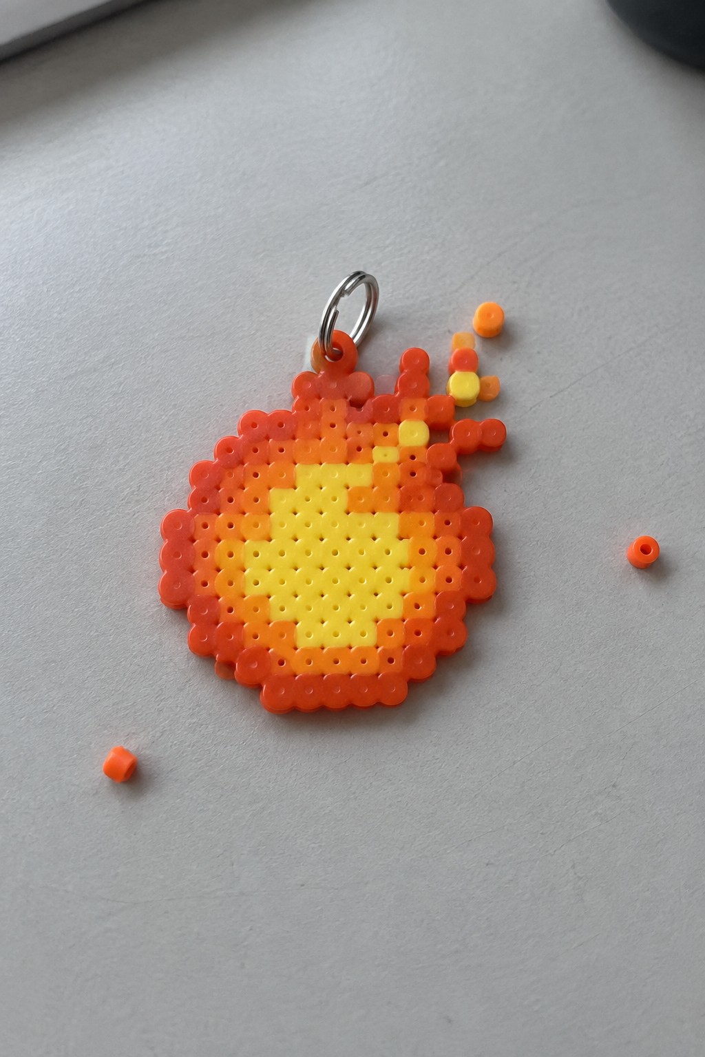 Perler bead fireball keychain in yellow, orange, and red on a pegboard with attached metal ring.