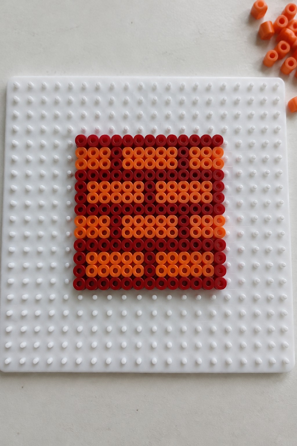 Red-orange gradient square of perler beads on white pegboard with loose beads nearby