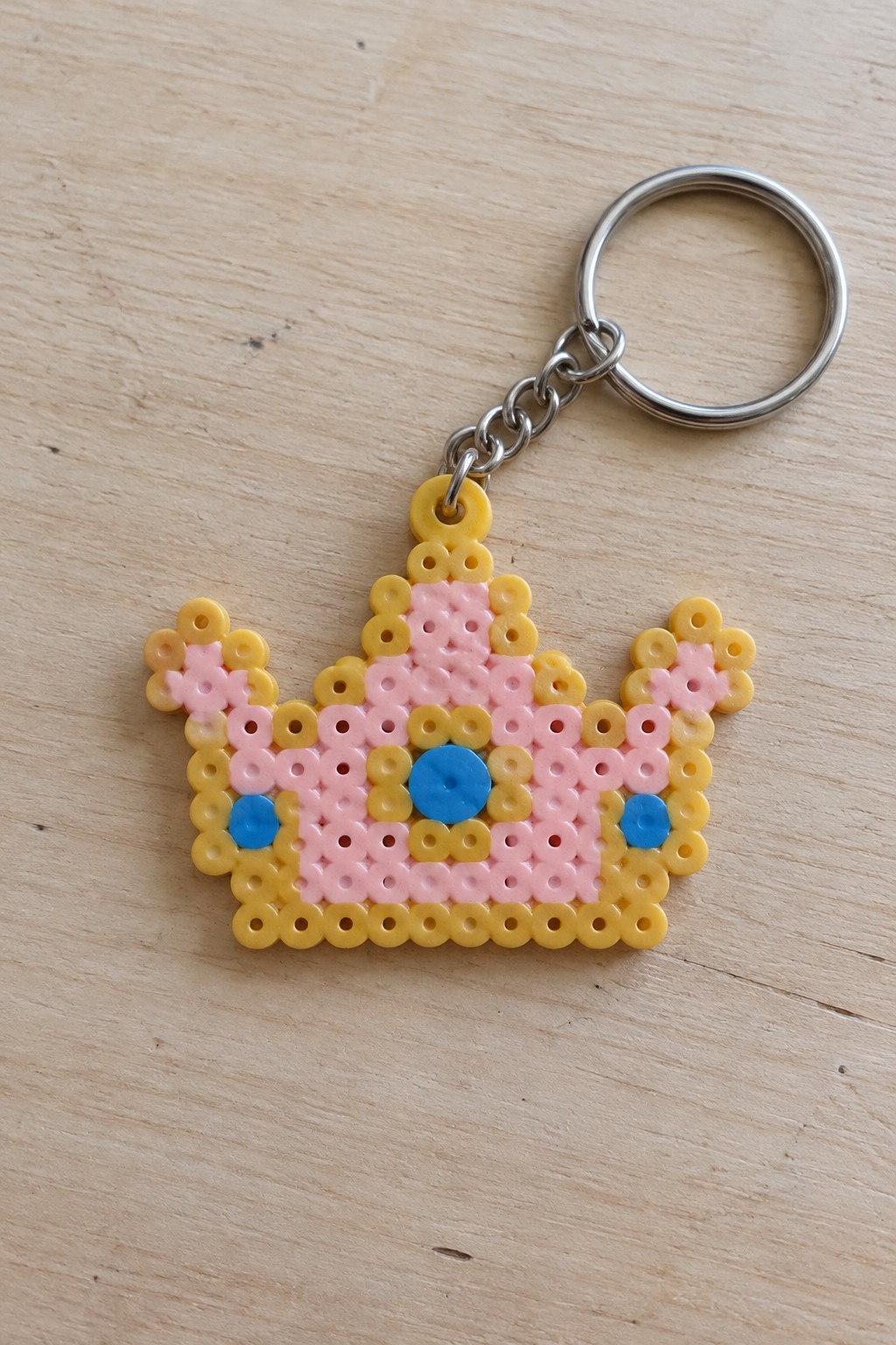 Pink and yellow perler bead crown keychain with blue accents on wood.