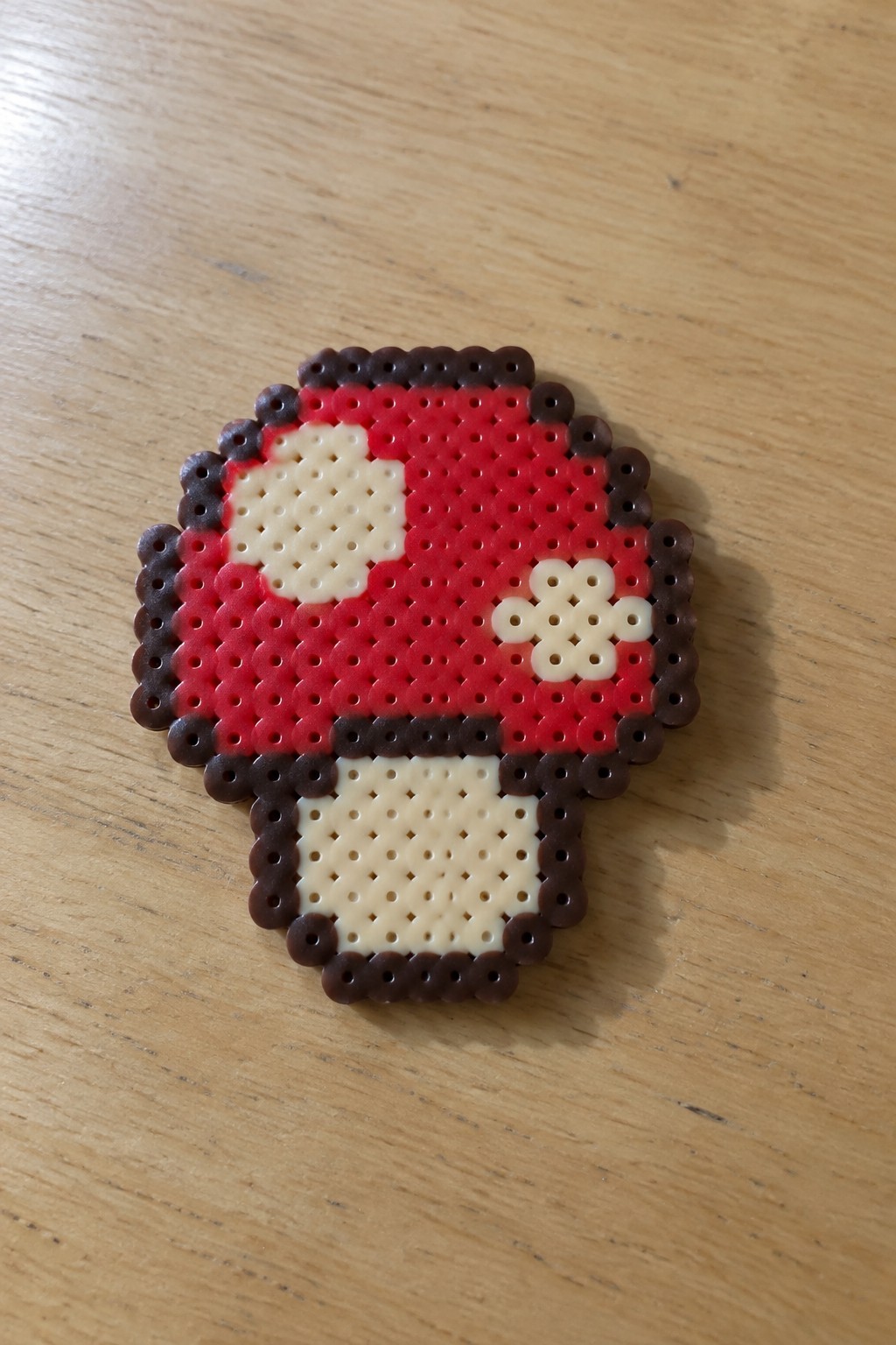 Red perler bead mushroom with white spots, brown border, and beige stem on wood.