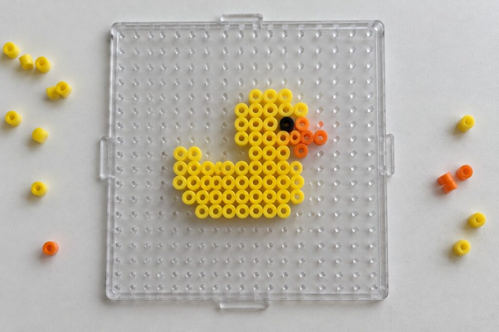 18 Easy Perler Bead Ideas Featuring Cute Mini Designs Yellow duck perler bead art on clear pegboard with black eye, orange beak, and loose beads nearby