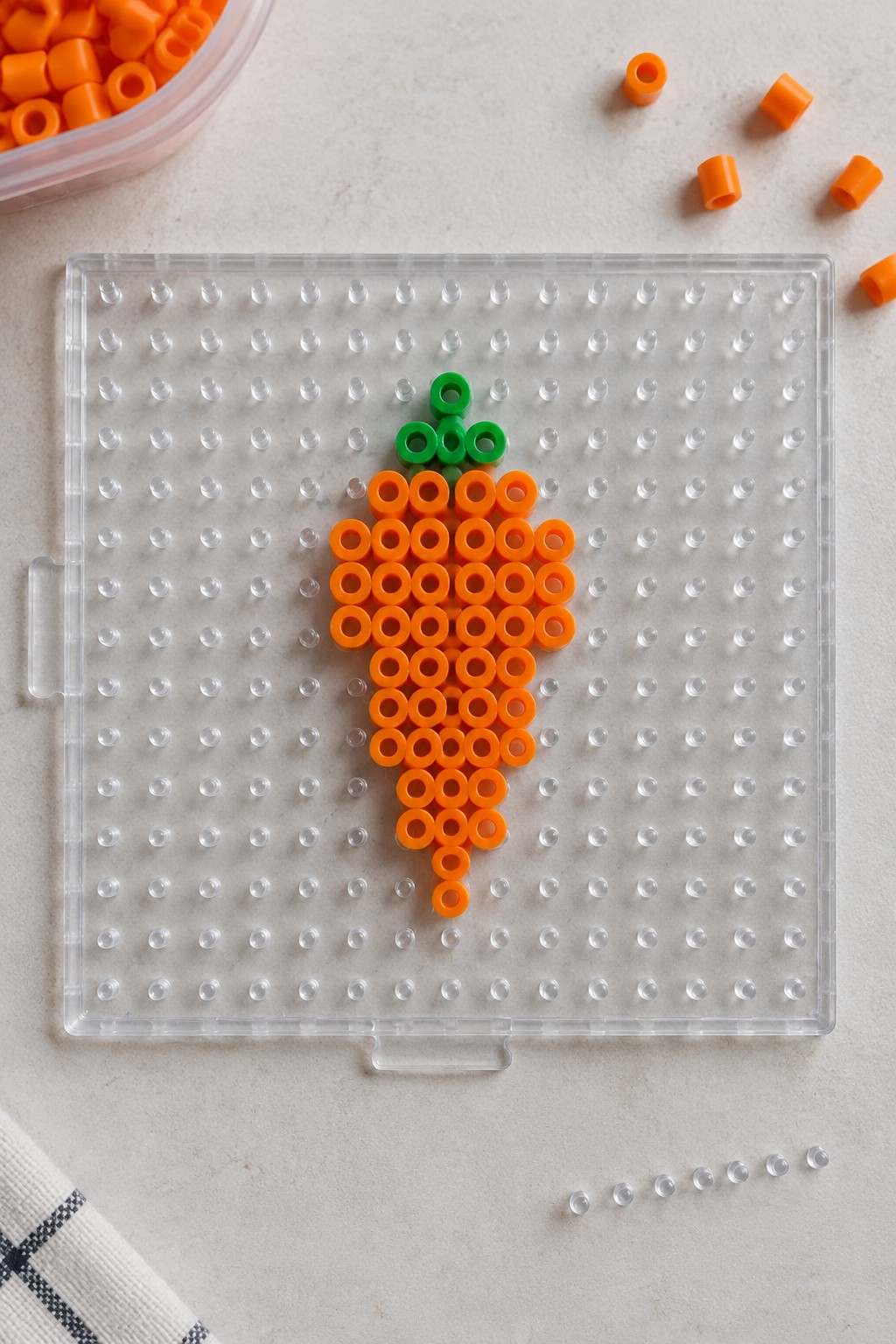 Perler bead carrot in orange and green on clear plastic pegboard