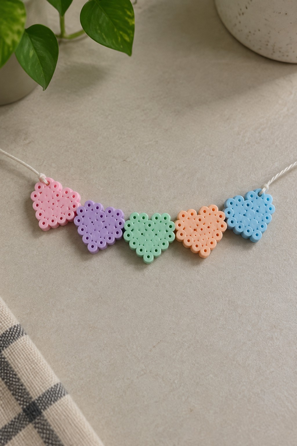 Five small fused Perler bead hearts in pink, purple, mint green, peach, and blue strung on white cord.