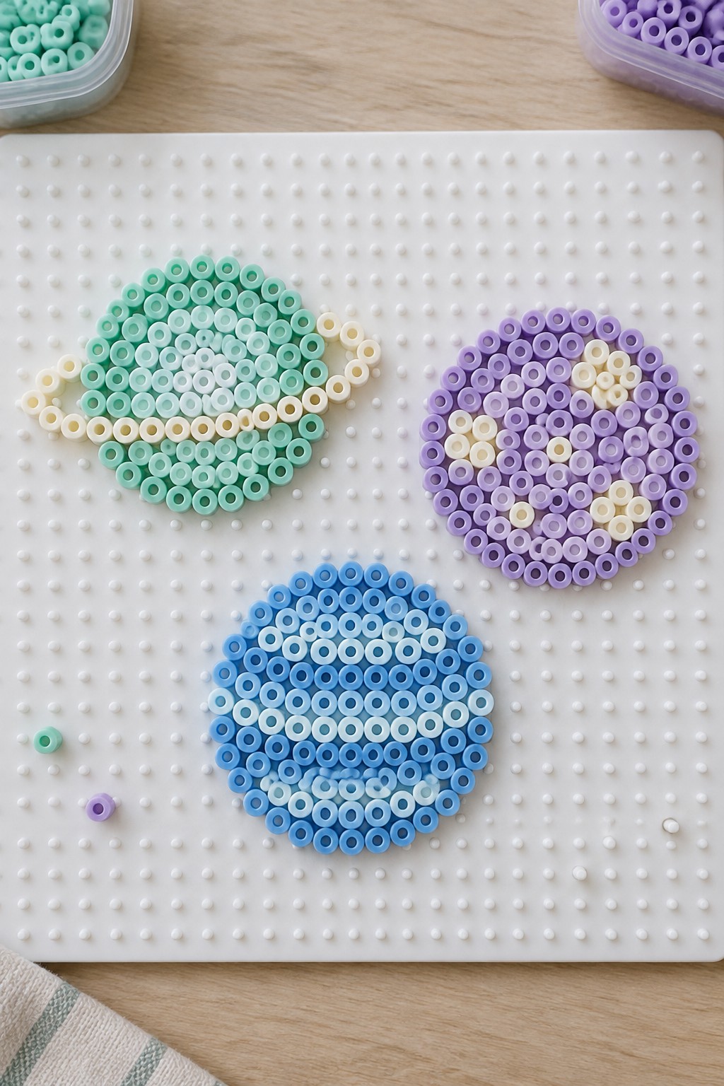Three unfused Perler bead planet designs on a pegboard showing a mint green ringed planet, a purple planet with highlights, and a blue gradient planet.