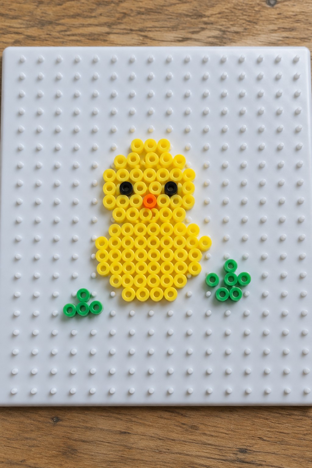 Yellow perler bead chick with black eyes, orange beak on white pegboard