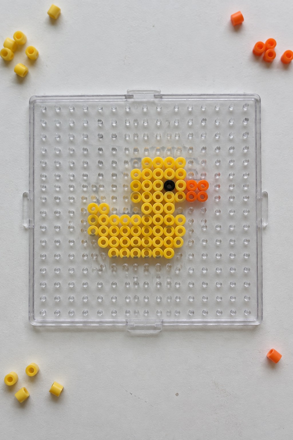 Yellow duck perler bead art on clear pegboard with black eye, orange beak, and loose beads nearby