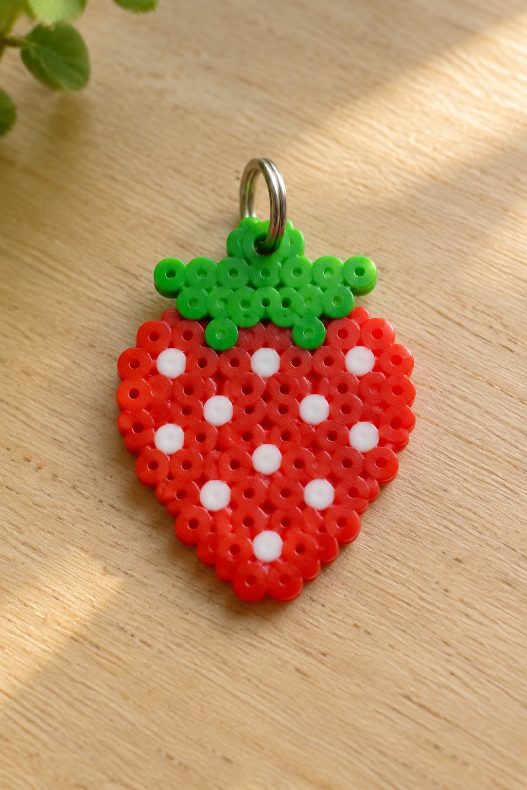 Perler bead strawberry keychain with red body, white dots, green top on wood.