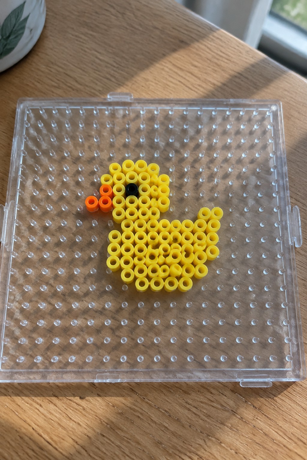 Yellow perler bead duck with orange beak and black eye on pegboard