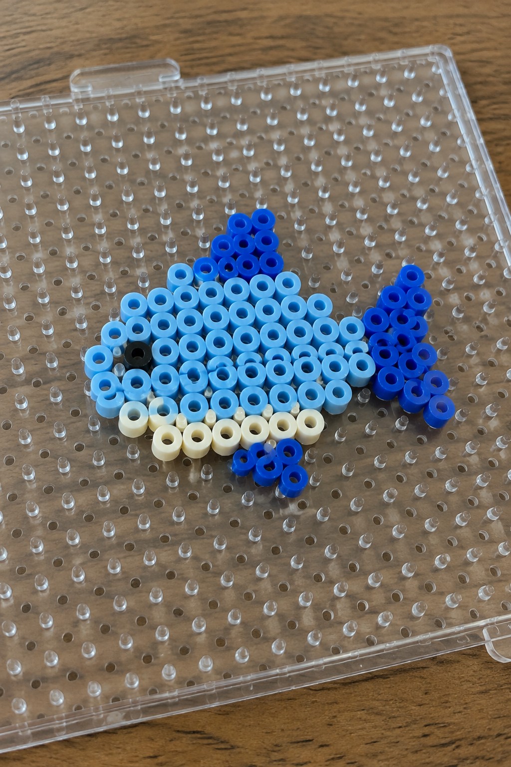 Blue perler bead fish with black eye on clear pegboard over wood