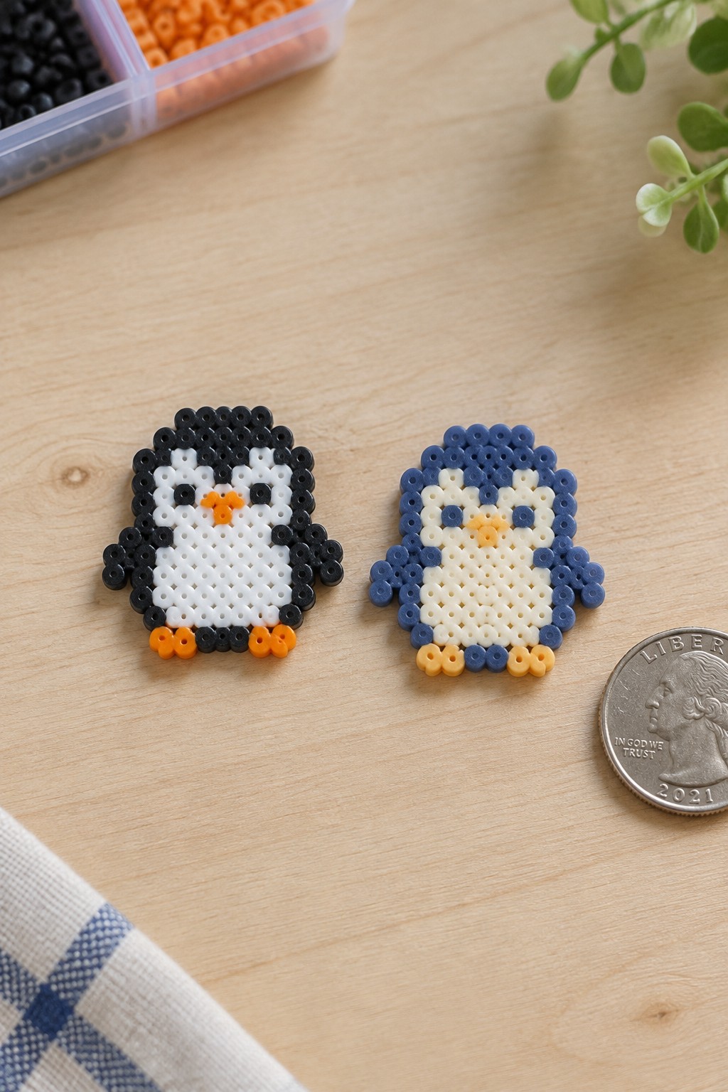 Two small fused Perler bead penguins, one black and white with orange beak and feet, the other blue and white with yellow beak and feet, next to a quarter.
