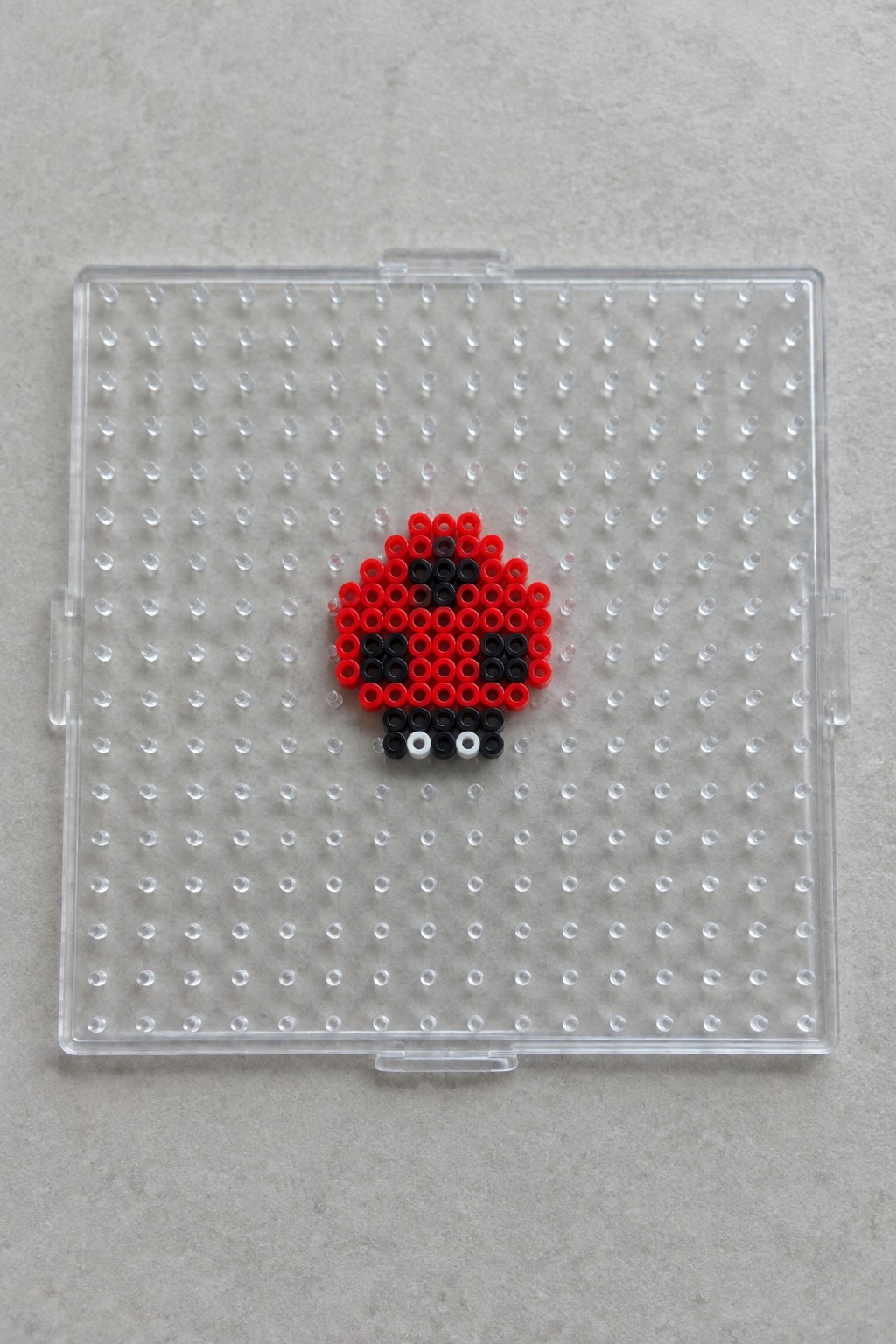 Red and black beaded ladybug with white eyes on clear pegboard.