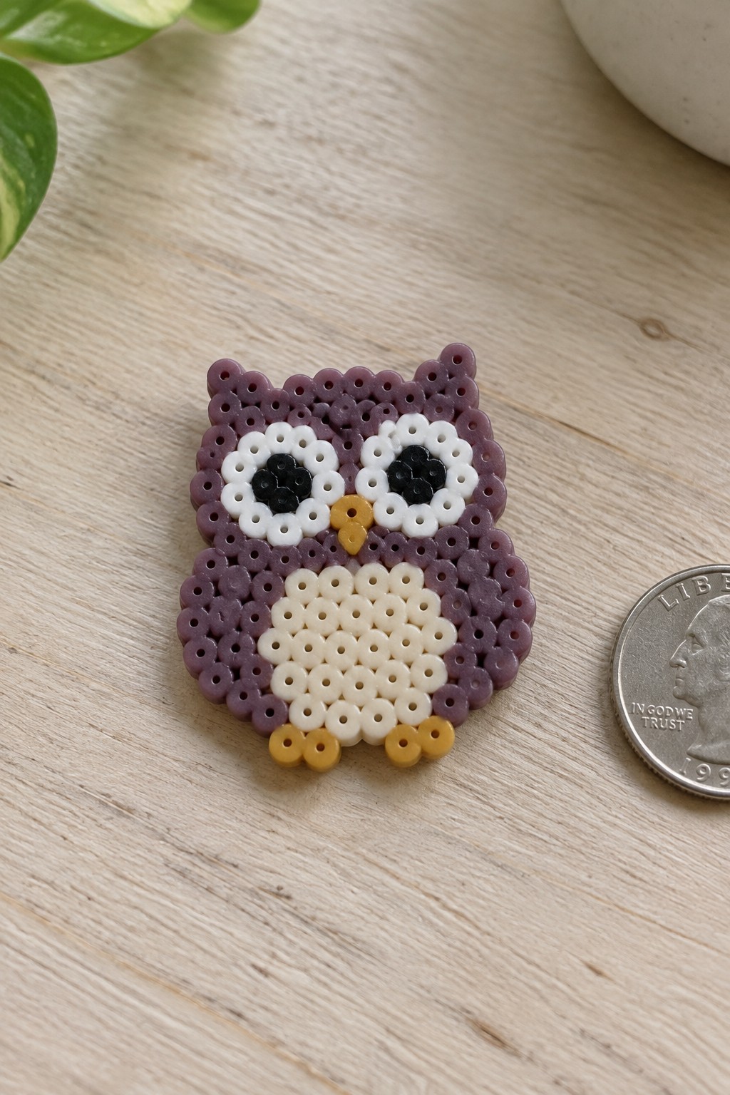 Purple perler bead owl with white belly beside US quarter on wood.