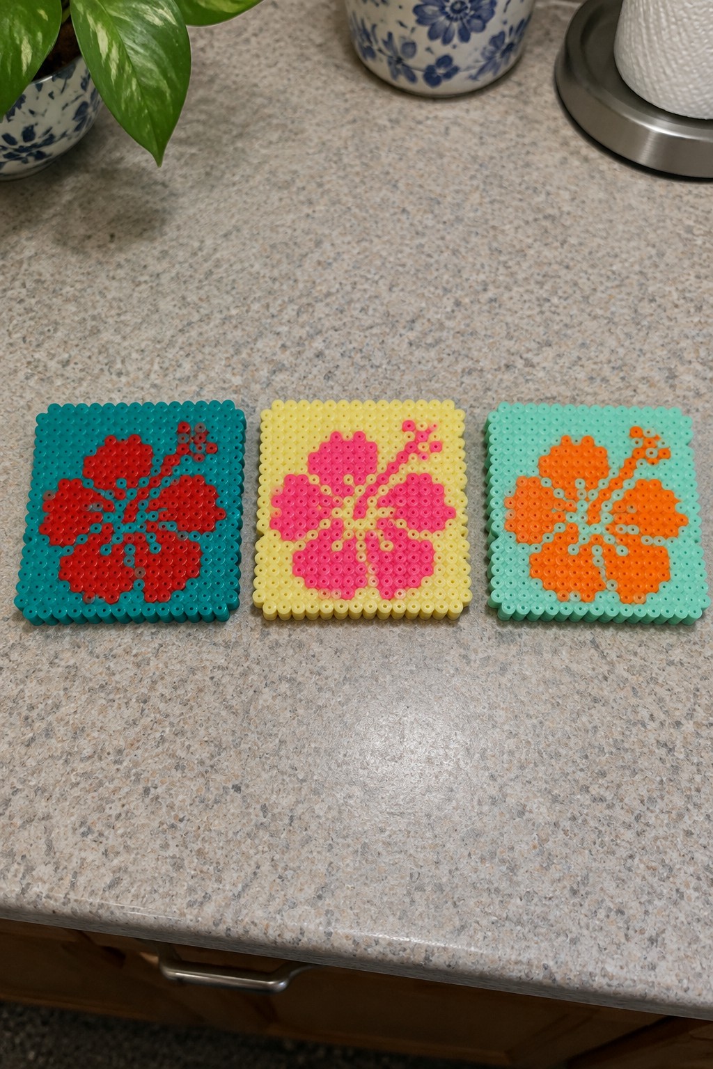 Three perler bead coasters with red, pink, and orange hibiscus flowers on a counter.