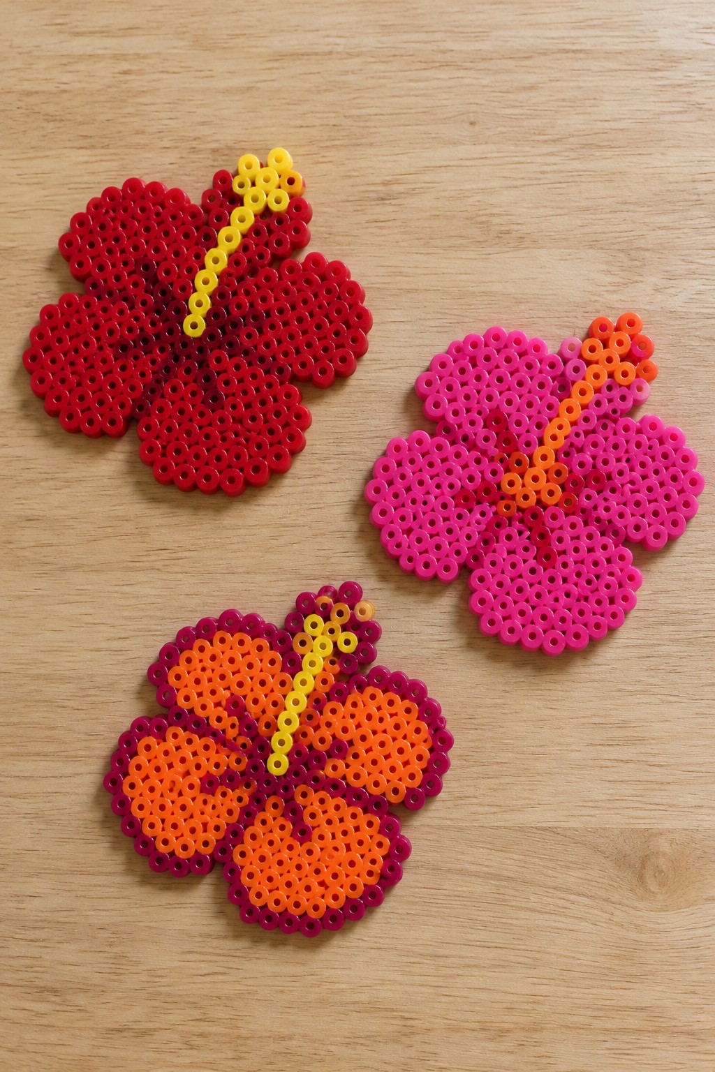 Three colorful perler bead hibiscus flowers on a wooden surface.