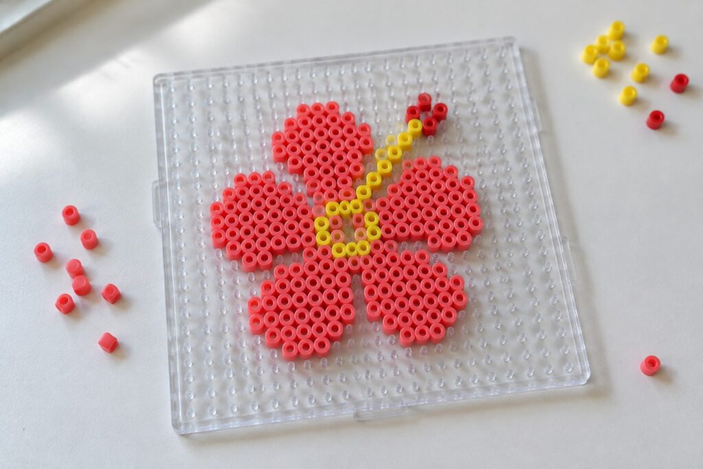 18 Perler Bead Flower Ideas Featuring Vibrant Hibiscus Blooms Clear pegboard with red perler bead flower, yellow center, and scattered loose beads