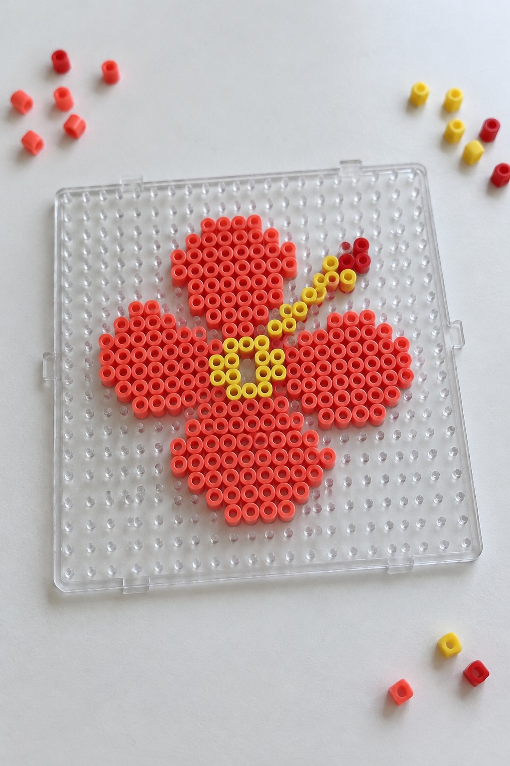 Clear pegboard with red perler bead flower, yellow center, and scattered loose beads