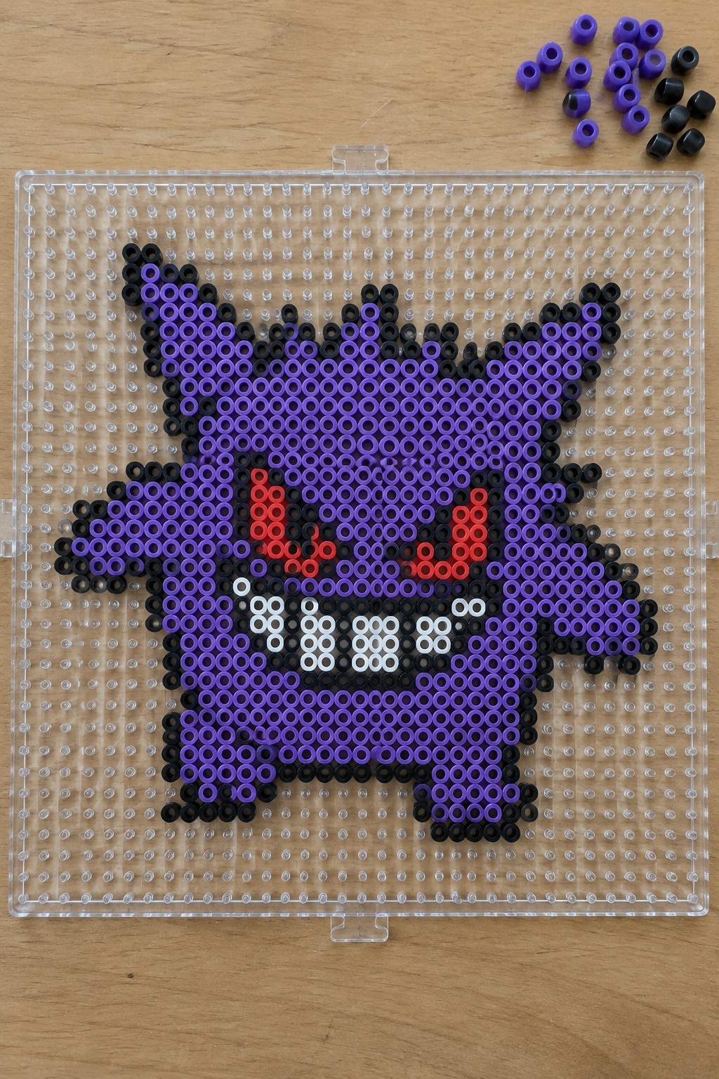 Purple black Gengar perler bead art on clear pegboard with loose beads nearby