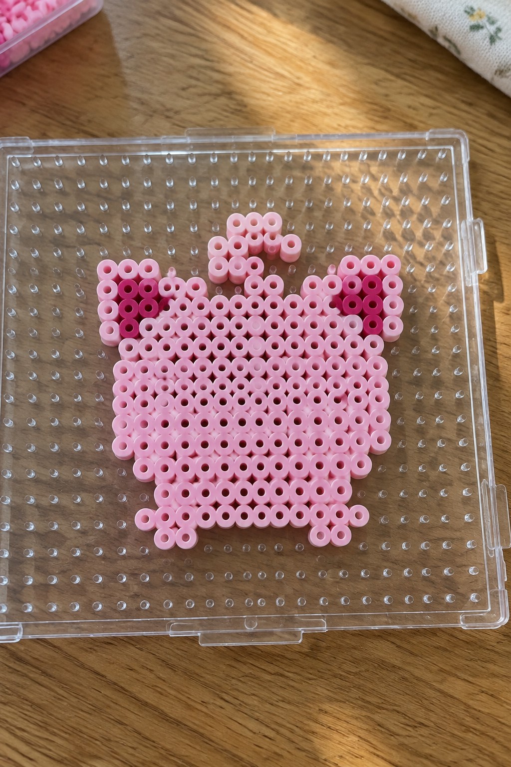 Pink perler beads forming a cat shape on clear pegboard over wood.