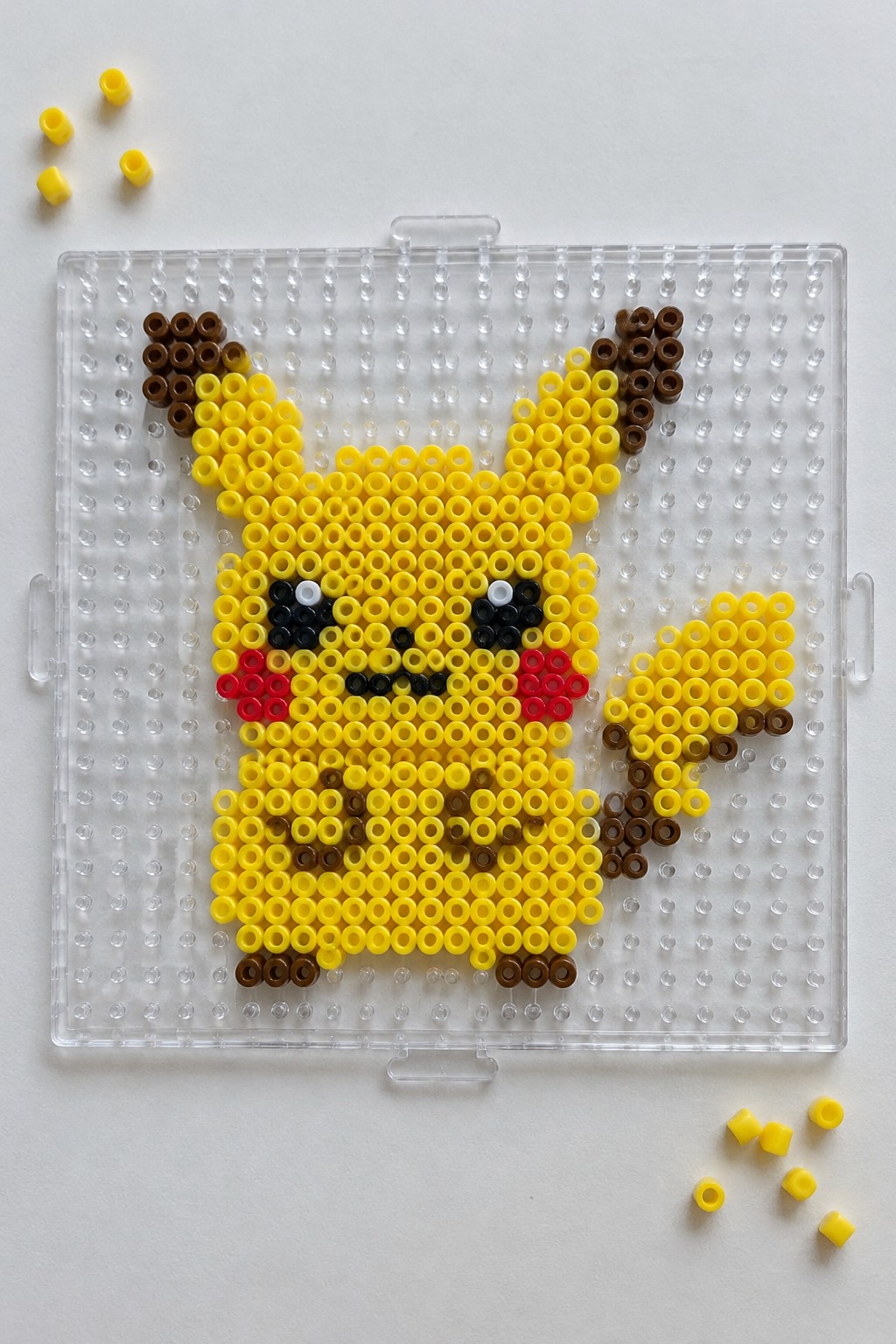 Yellow Pikachu Perler bead figure on clear pegboard with loose beads nearby