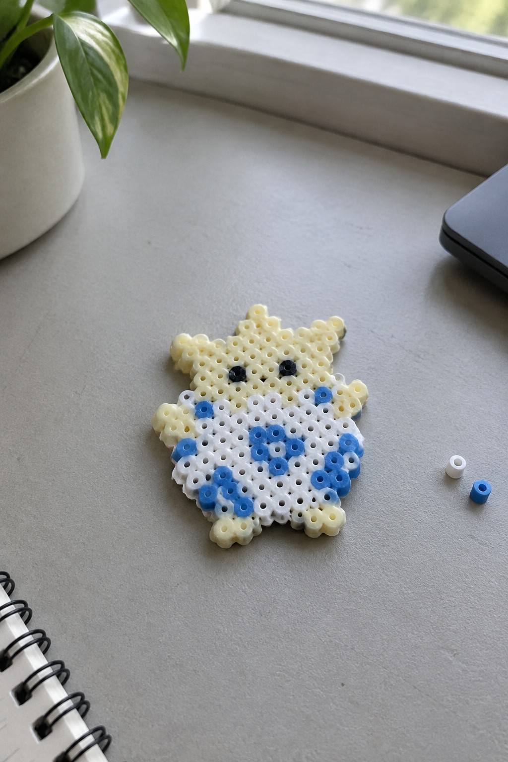 Yellow perler bead creature with black eyes, white body and blue patterns on desk