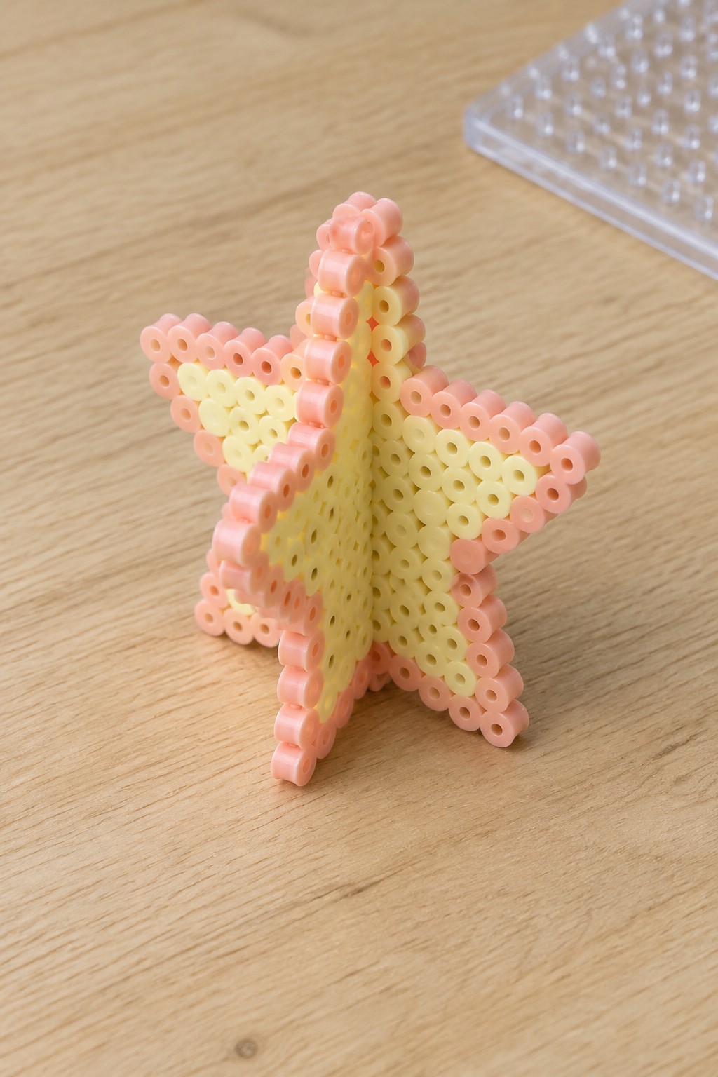 Pink and yellow Perler bead star upright on wooden table with pegboard nearby