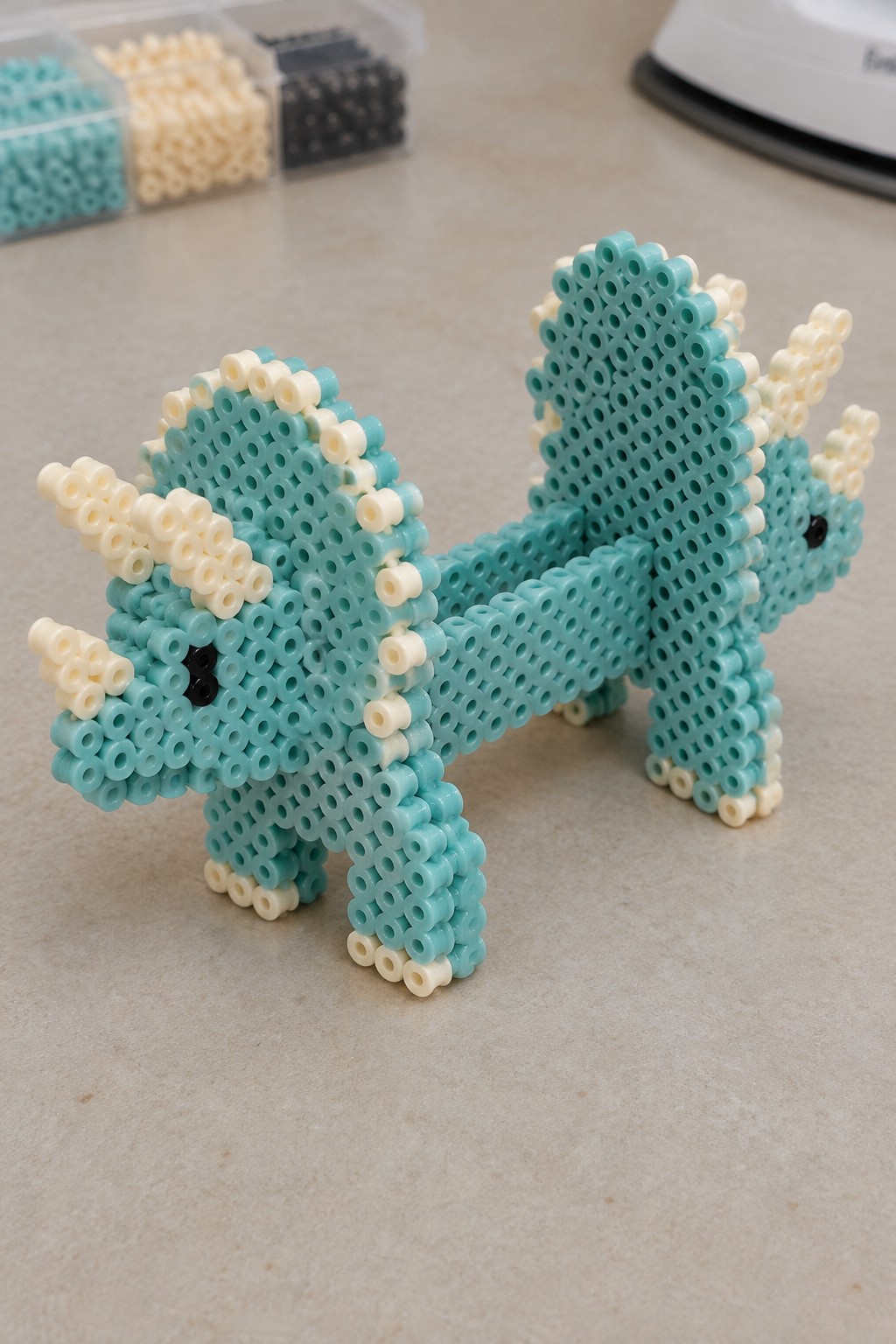 Turquoise perler bead dragon with cream horns and black eyes on table