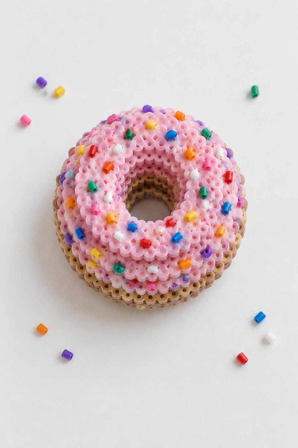 Pink perler bead donut with colorful sprinkles on white surface with loose beads.