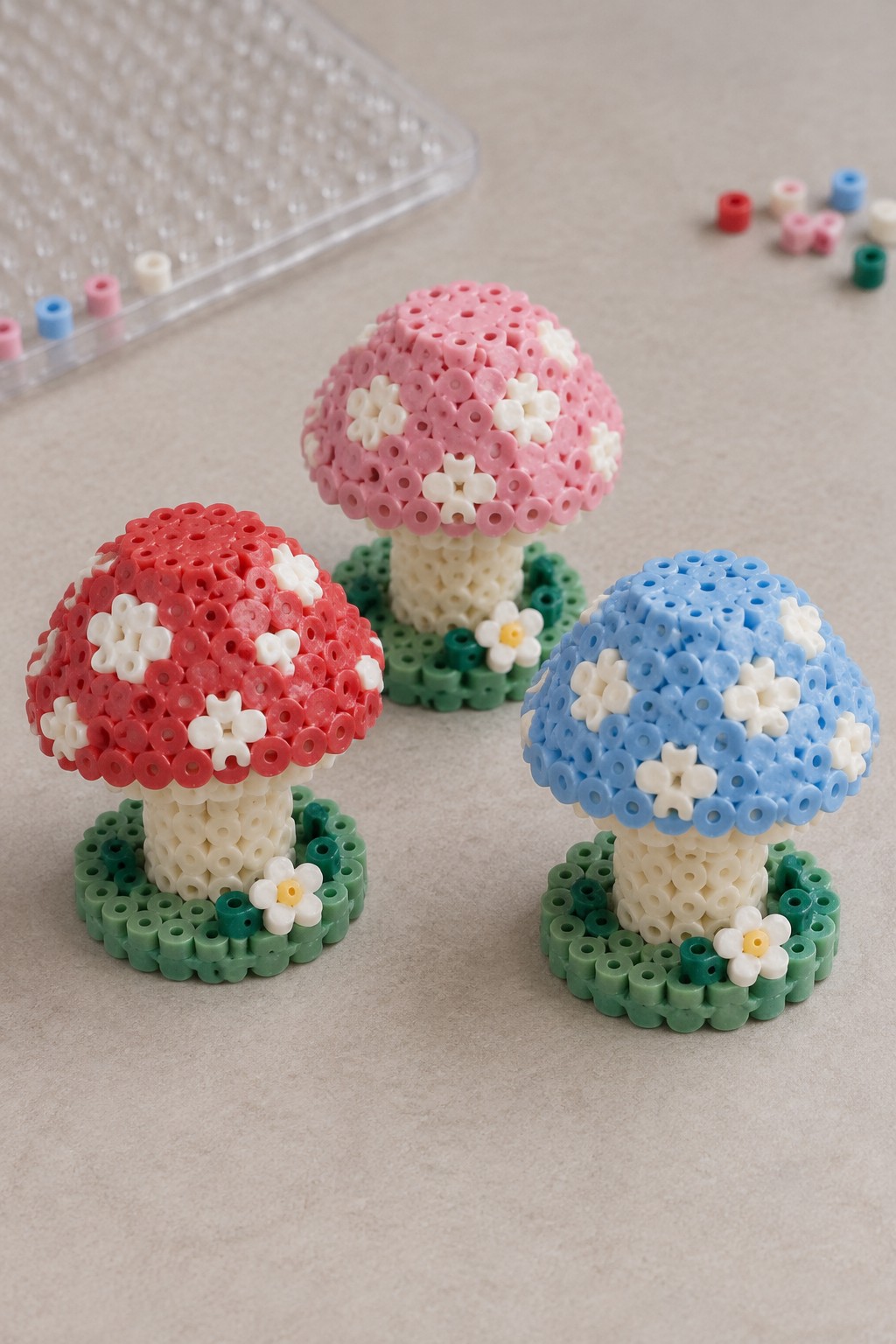 Three small 3D Perler bead mushrooms in red, pink, and blue with white spots, each on a green base with a flower accent.