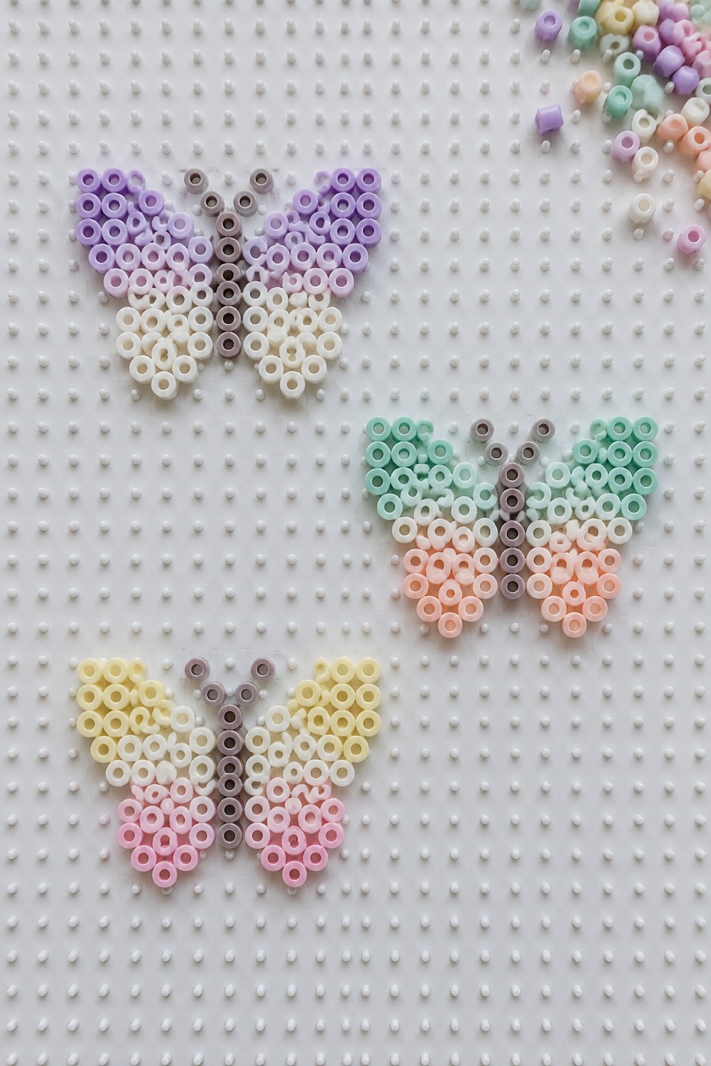 Three pastel gradient perler bead butterflies on a white pegboard with loose beads.