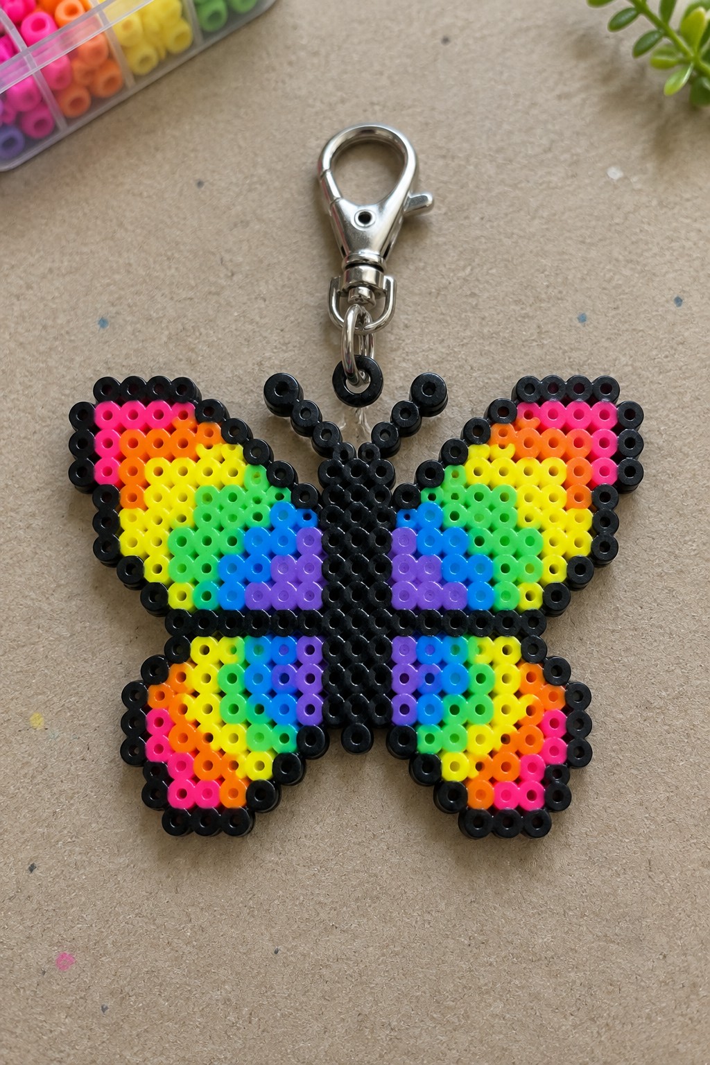 Rainbow perler bead butterfly keychain with silver clasp on beige surface.