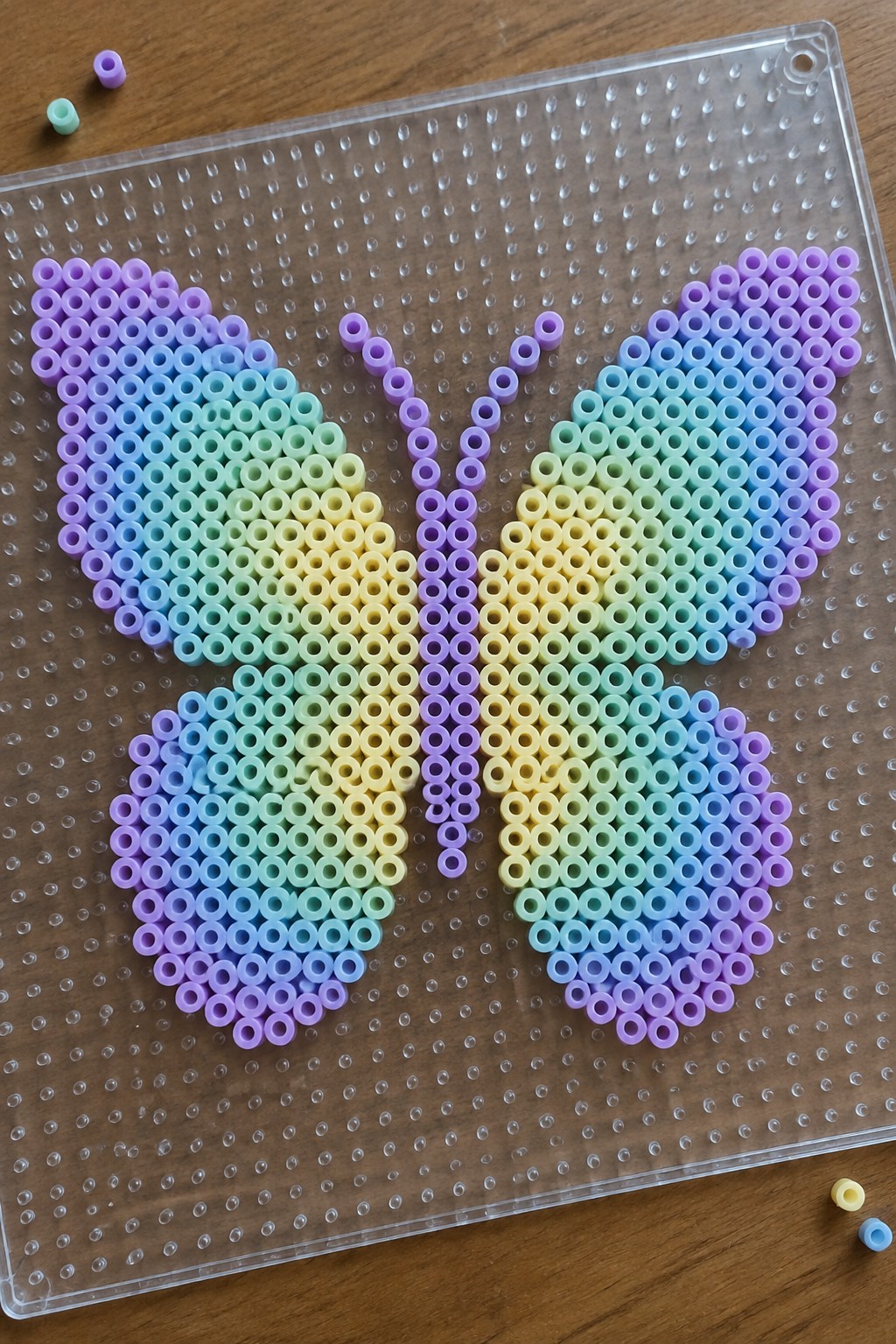 Rainbow perler bead butterfly with gradient colors on clear pegboard over wood