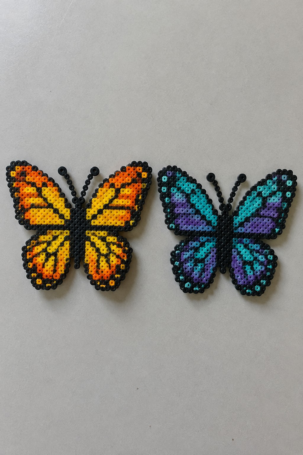 Two Perler bead butterflies, orange-yellow and blue-purple, on light gray surface.