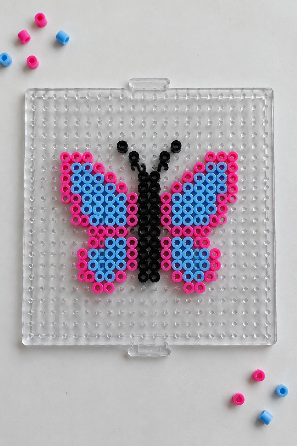 Perler bead butterfly with pink and blue symmetrical wings and black body on a pegboard.