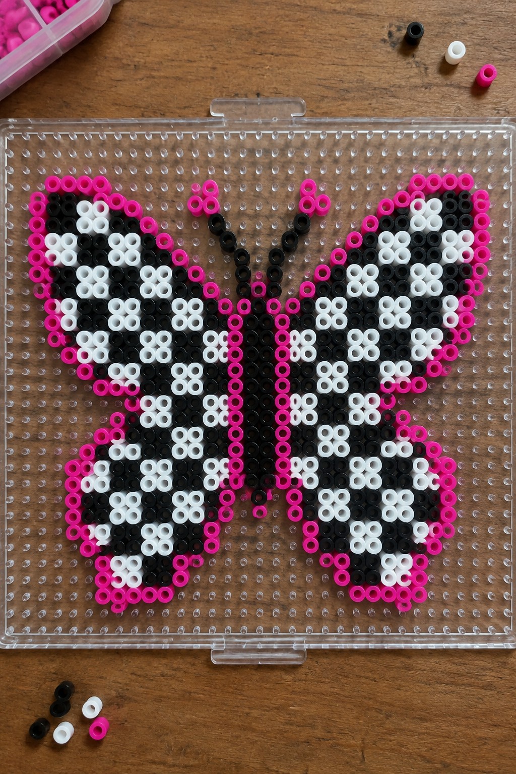 Pink black and white perler bead butterfly on clear pegboard with loose beads