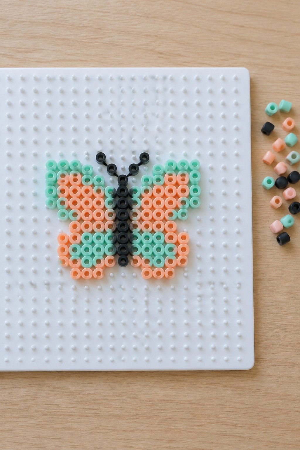 Perler bead butterfly with peach and mint wings on white pegboard beside loose beads
