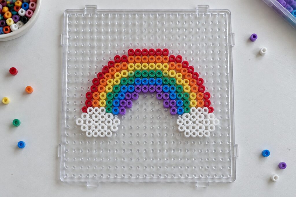 19 Colorful Rainbow Perler Bead Designs for Beginner Crafters Rainbow Perler bead design on clear pegboard with loose colorful beads nearby