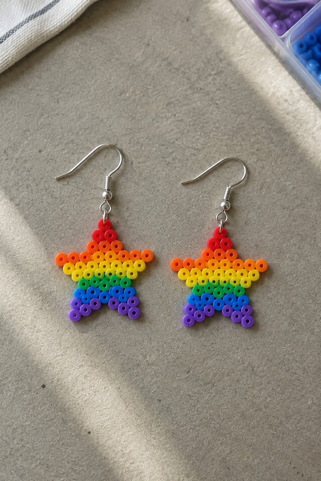 Two rainbow beaded star earrings with silver hooks on beige surface