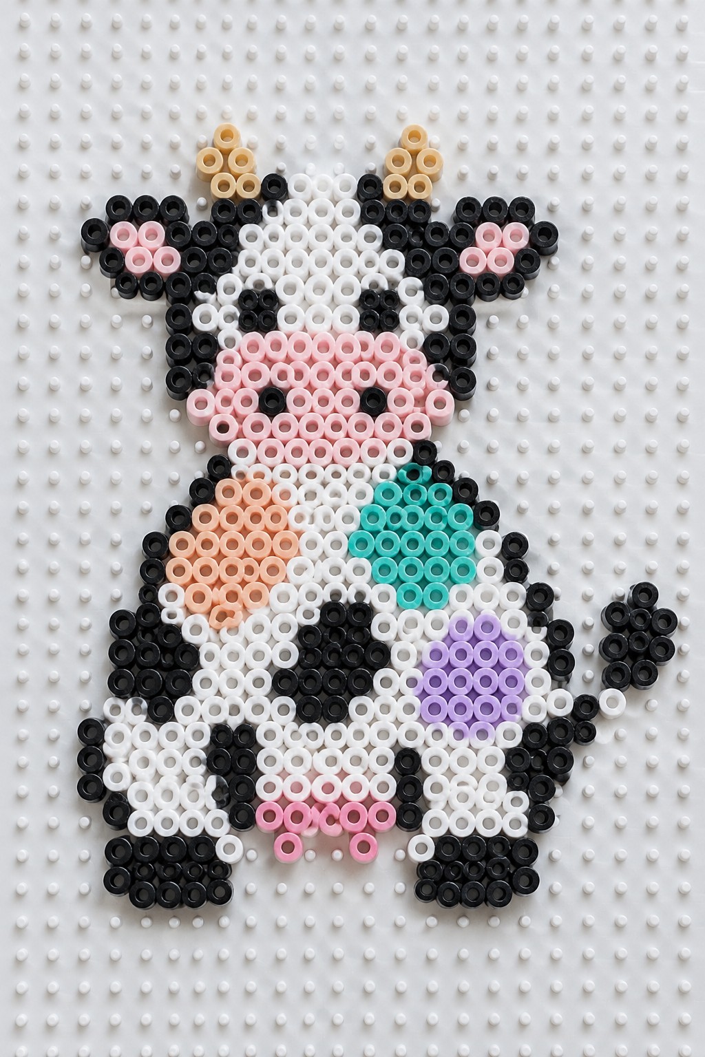 Perler bead cow in black, white, and pink with teal accents on pegboard.