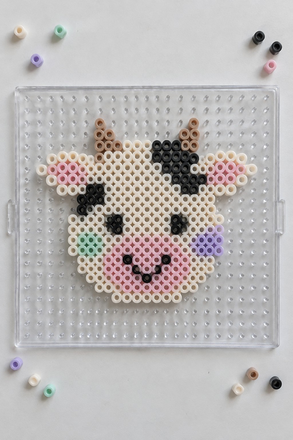 Perler bead cow face on clear pegboard with scattered loose beads nearby