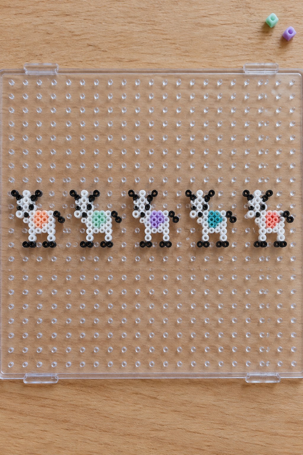 Five perler bead goats with pastel patches on a clear pegboard over wood.