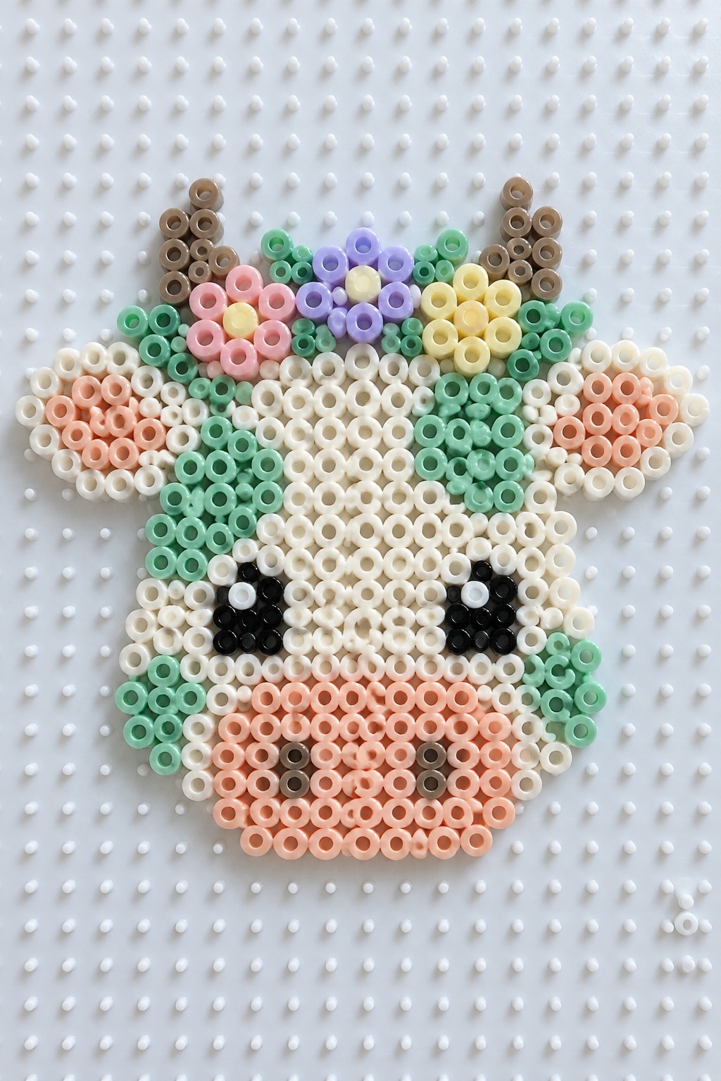 Perler bead cow face with pastel flower crown, mint green spots, and peach ears arranged on a white pegboard.
