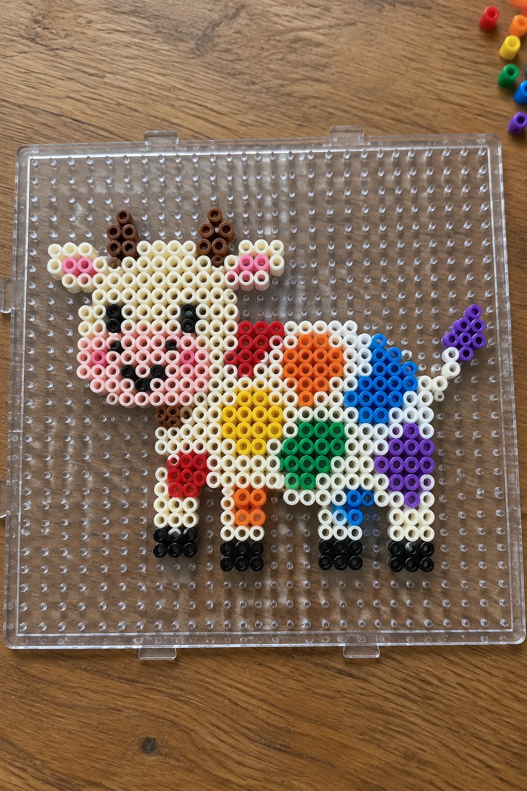 Perler bead cow with rainbow stripes on its body, brown horns, pink ears, and black hooves arranged on a clear pegboard.