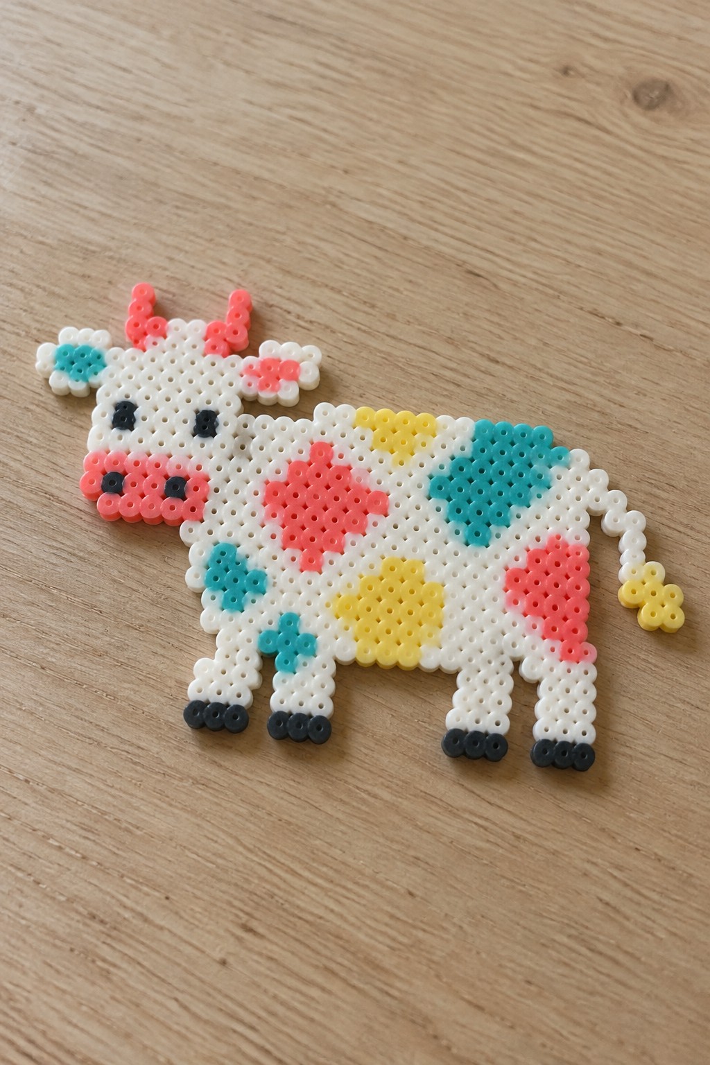 Colorful perler bead cow with pink, yellow, and cyan spots on wood