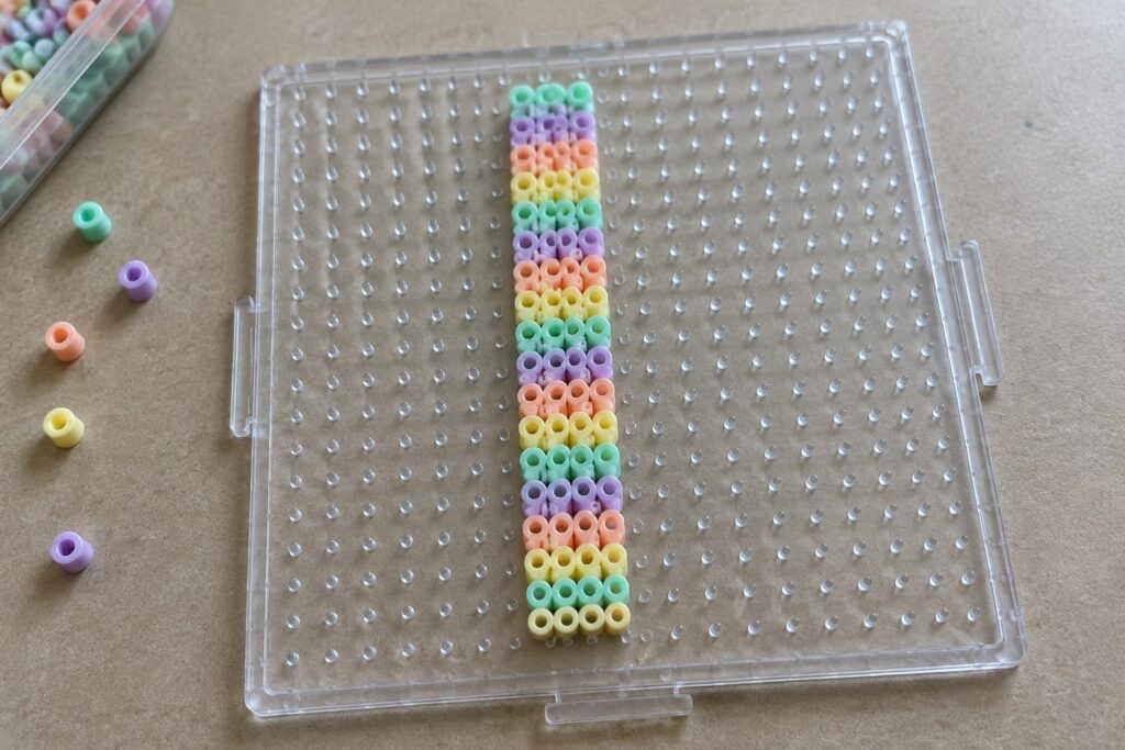 19 Creative Perler Bead Bookmarks Using Slim Patterns Clear pegboard with vertical pastel beads in rainbow stripes and scattered loose beads.