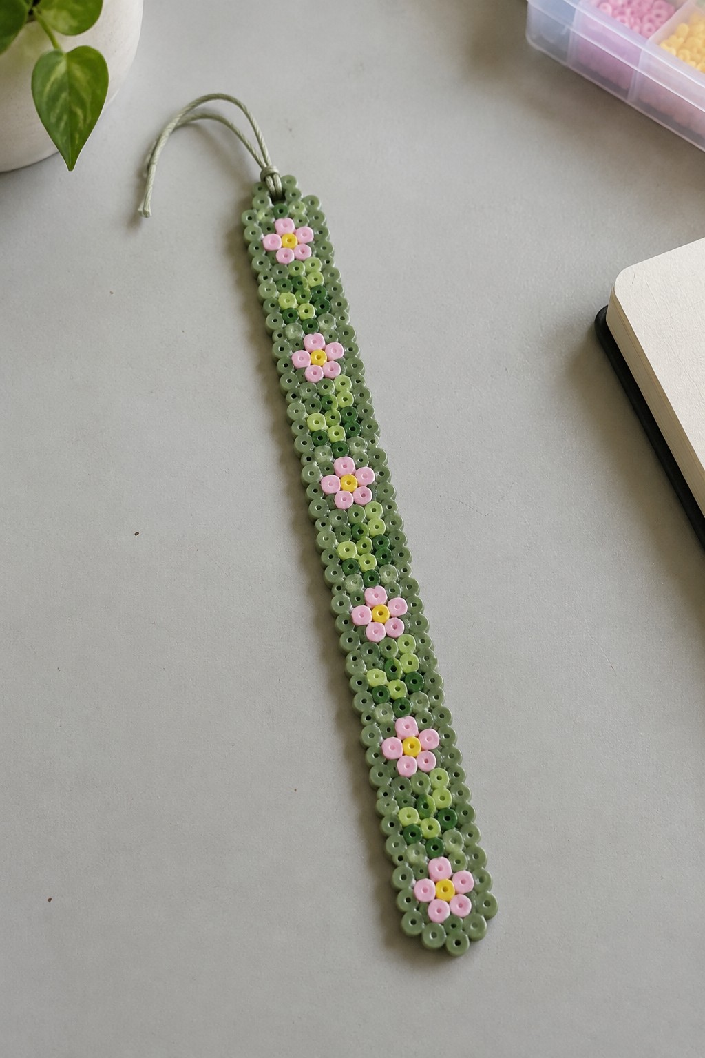 A slim Perler bead bookmark made of green beads with pink flowers that have yellow centers.