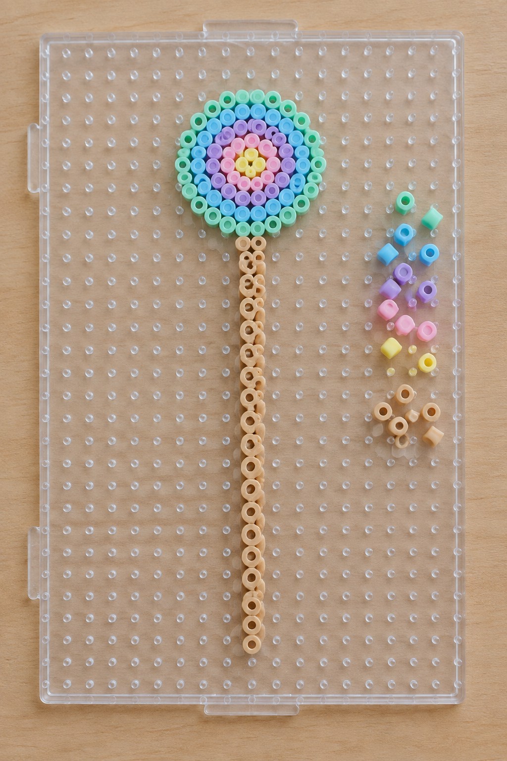 Perler bead rainbow lollipop with beige stem on clear pegboard beside loose beads