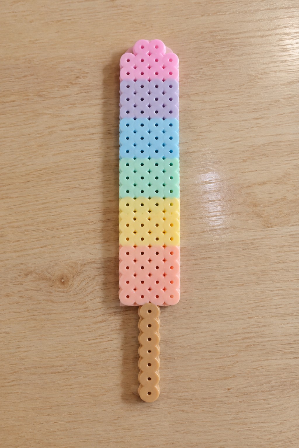 Rainbow perler bead popsicle with pink-to-peach stripes and beige stick on wood.