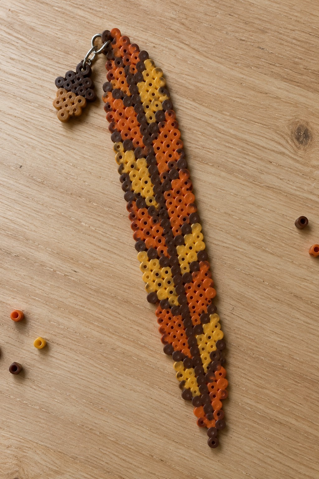 Fused Perler bead bookmark in a slim tapered leaf shape with brown, orange, and yellow diagonal color blocks, attached to a metal clasp with a small beaded charm.