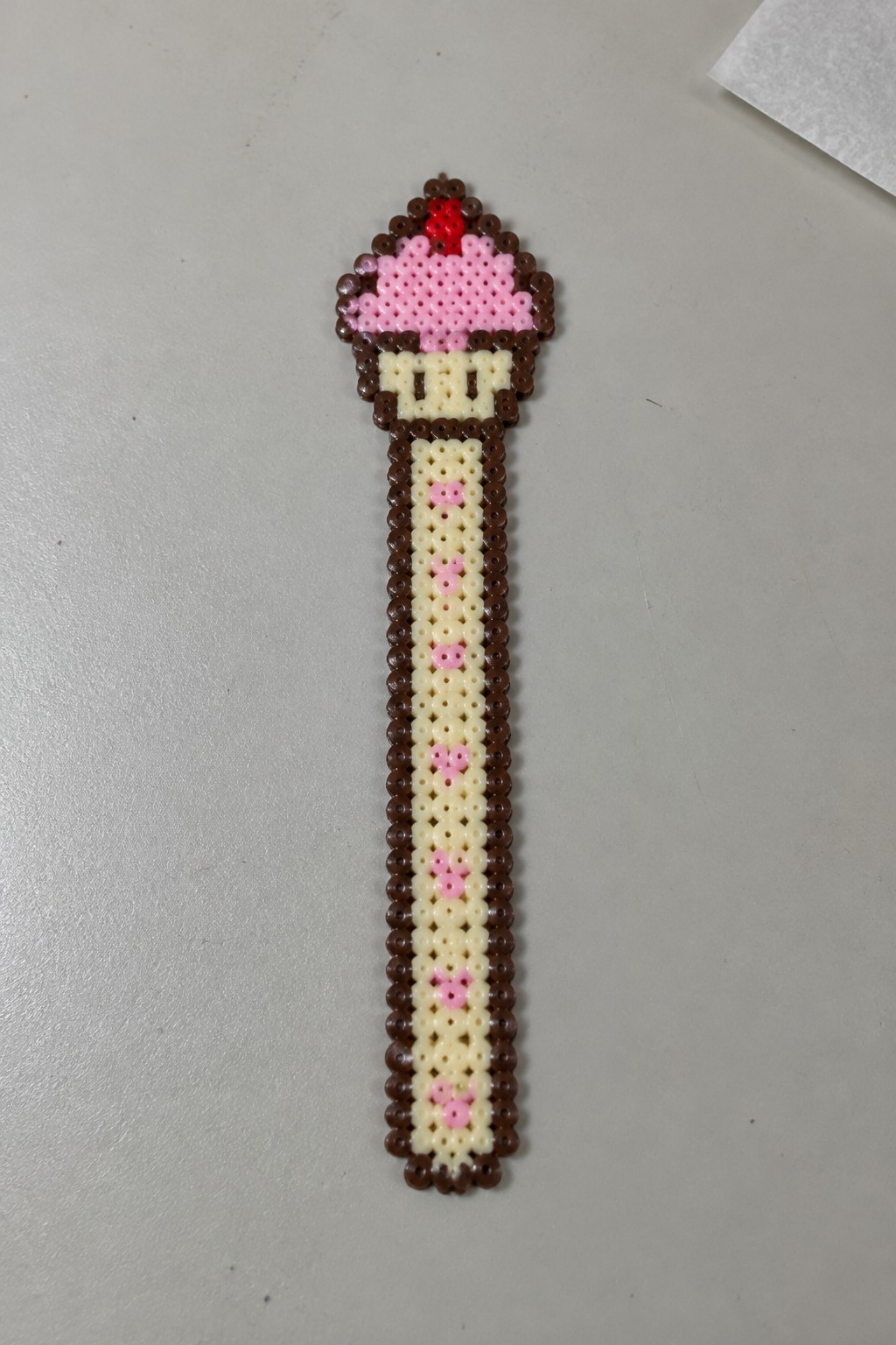 Perler bead cupcake on a stick with pink hearts on cream and brown handle.