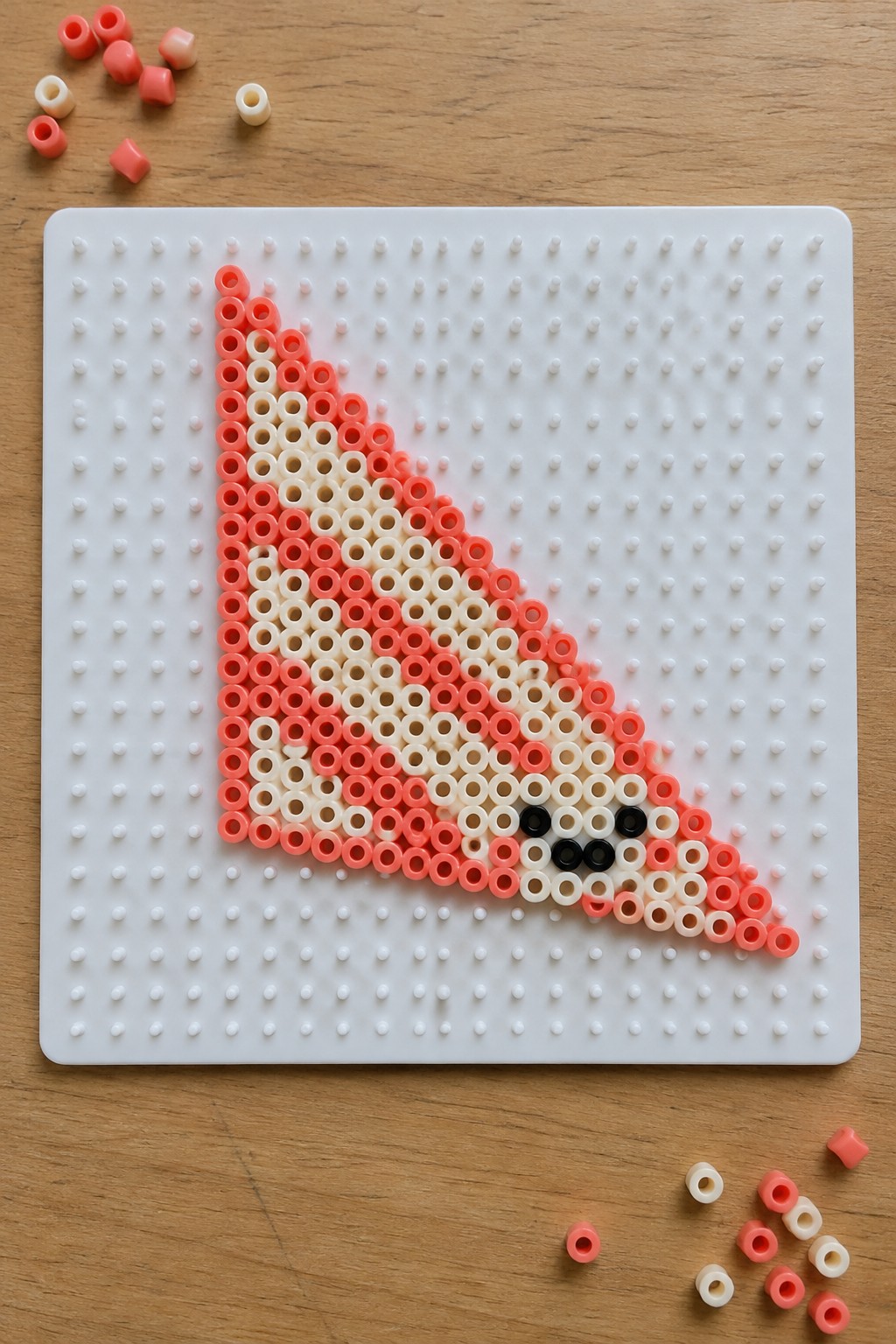 Pink and white perler beads forming a shark on pegboard with loose beads on wood.